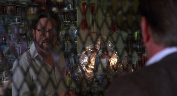 Movie still from “The Believers” (1987), directed by John Schlesinger – A man sitting at a table in front of a metal fence; Medium shot, Over the shoulder angle