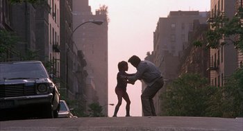 Movie still from “The Believers” (1987), directed by John Schlesinger – A man and a girl are dancing on the street; Wide shot, Low angle