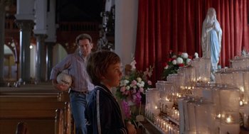 Movie still from “The Believers” (1987), directed by John Schlesinger – A man and a boy standing in front of a wall of lit candles; Medium shot, Over the shoulder angle