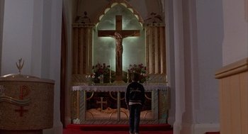 Movie still from “The Believers” (1987), directed by John Schlesinger – A person standing in front of a crucifix in a church; Wide shot, Low angle