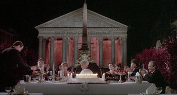 Movie still from “The Belly of an Architect” (1987), directed by Peter Greenaway – A group of people sitting around a table with a cake on it; Wide shot, Low angle