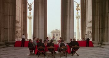 Movie still from “The Belly of an Architect” (1987), directed by Peter Greenaway – A group of people sitting around a table in front of columns; Extreme Wide shot, High angle