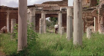 Movie still from “The Belly of an Architect” (1987), directed by Peter Greenaway – A man walking through a field of grass near some pillars; Extreme Wide shot, Low angle