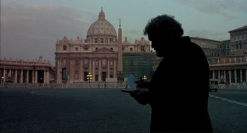 Movie still from “The Belly of an Architect” (1987), directed by Peter Greenaway – A man standing in front of a building looking at his cell phone; Wide shot, Low angle