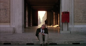 Movie still from “The Belly of an Architect” (1987), directed by Peter Greenaway – A man sitting on the steps of a building looking at his cell phone; Wide shot, Low angle