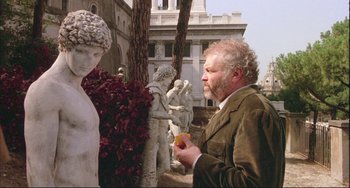 Movie still from “The Belly of an Architect” (1987), directed by Peter Greenaway – A man standing in front of a statue holding something; Medium shot, Over the shoulder angle