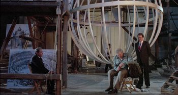 Movie still from “The Belly of an Architect” (1987), directed by Peter Greenaway – An older man sitting in a chair next to a woman; Wide shot, Low angle