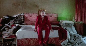 Movie still from “The Belly of an Architect” (1987), directed by Peter Greenaway – A man sitting on top of a bed wearing a red suit; Wide shot, Low angle