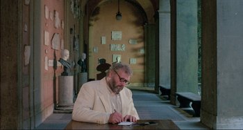 Movie still from “The Belly of an Architect” (1987), directed by Peter Greenaway – A man sitting at a table writing on a piece of paper; Wide shot, High angle