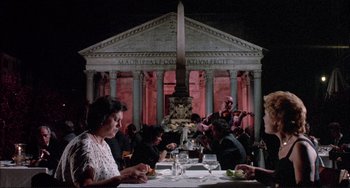 Movie still from “The Belly of an Architect” (1987), directed by Peter Greenaway – A group of people sitting at a table in front of a building; Wide shot, Low angle