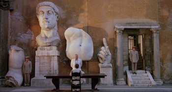 Movie still from “The Belly of an Architect” (1987), directed by Peter Greenaway – A man sitting at a table in front of a statue; Extreme Wide shot, Low angle