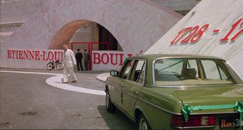 Movie still from “The Belly of an Architect” (1987), directed by Peter Greenaway – A green car parked in front of a building with people walking around; Wide shot, Over the shoulder angle