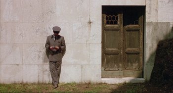 Movie still from “The Belly of an Architect” (1987), directed by Peter Greenaway – A man standing in front of a building reading a book; Wide shot, Low angle