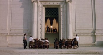 Movie still from “The Belly of an Architect” (1987), directed by Peter Greenaway – A group of people sitting in front of an entrance to a building; Extreme Wide shot, Low angle