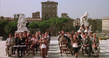 Movie still from “The Belly of an Architect” (1987), directed by Peter Greenaway – A group of people sitting in chairs in front of a building; Extreme Wide shot, High angle