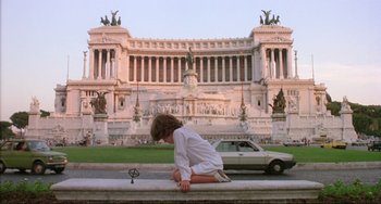 Movie still from “The Belly of an Architect” (1987), directed by Peter Greenaway – A person sitting on a ledge in front of a building; Extreme Wide shot, Low angle