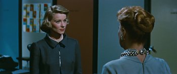 Movie still from “The Best of Everything” (1959), directed by Jean Negulesco – A woman in a black dress and pearls looking at a mirror; Medium shot, Over the shoulder angle