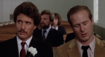 Movie still from “The Big Chill” (1983), directed by Lawrence Kasdan – Two men in suits and ties sitting in front of other men; Close Up shot, Over the shoulder angle