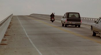 Movie still from “The Big Chill” (1983), directed by Lawrence Kasdan – A person riding a motorcycle on the side of a road; Wide shot, High angle