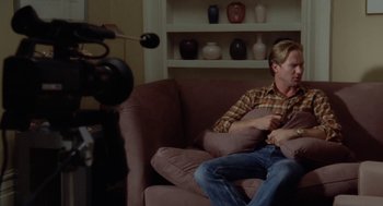 Movie still from “The Big Chill” (1983), directed by Lawrence Kasdan – A man sitting on top of a couch in a living room; Medium shot, Over the shoulder angle