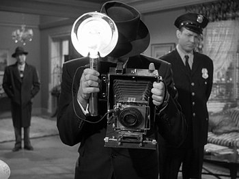 Movie still from “The Big Heat” (1953), directed by Fritz Lang – A man in a suit and hat holding an old camera; Medium shot, Over the shoulder angle