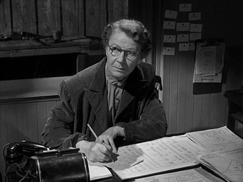 Movie still from “The Big Heat” (1953), directed by Fritz Lang – An older woman sitting at a desk writing on a piece of paper; Medium shot, Low angle