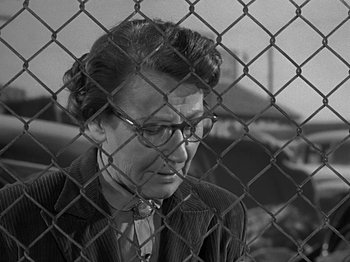 Movie still from “The Big Heat” (1953), directed by Fritz Lang – An older woman wearing a suit and glasses is behind a chain link fence; Close Up shot, Over the shoulder angle