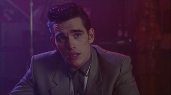 Movie still from “The Big Town” (1987), directed by Harold Becker – A man wearing a suit and tie sitting in front of a purple background; Close Up shot, Over the shoulder angle