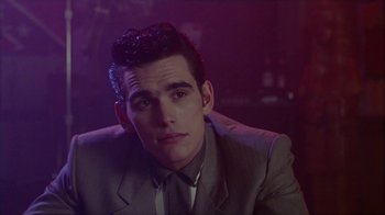 Movie still from “The Big Town” (1987), directed by Harold Becker – A man wearing a suit and tie sitting in front of a purple background; Close Up shot, Over the shoulder angle