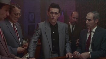 Movie still from “The Big Town” (1987), directed by Harold Becker – A man in a suit and tie standing next to a table; Medium shot, Low angle