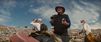 Movie still from “The Big Year” (2011), directed by David Frankel – A man in a hat is standing in a pile of trash; Medium shot, Low angle