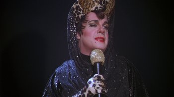 Movie still from “The Birdcage” (1996), directed by Mike Nichols – A woman in a leopard print hat holding a microphone; Close Up shot, Over the shoulder angle