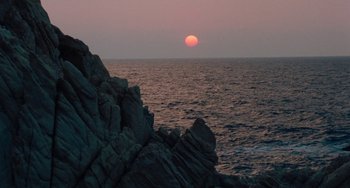 Movie still from “The Black Stallion” (1979), directed by Carroll Ballard – The sun is setting over the ocean with a large rock in the foreground; Extreme Wide shot, High angle