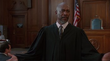 Movie still from “The Bonfire of the Vanities” (1990), directed by Brian De Palma – A man in a robe and tie in front of a flag; Medium shot, Low angle