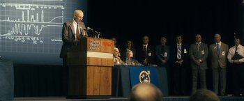 Movie still from “The Box” (2009), directed by Richard Kelly – A man is speaking at a podium in front of a group of people; Medium shot, Low angle