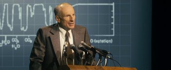 Movie still from “The Box” (2009), directed by Richard Kelly – A man in a suit and tie speaking into microphones in front of microphones; Medium shot, Over the shoulder angle