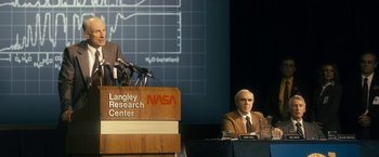 Movie still from “The Box” (2009), directed by Richard Kelly – A man in a suit and tie sitting at a podium in front of microphones; Medium shot, Low angle