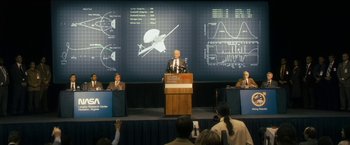 Movie still from “The Box” (2009), directed by Richard Kelly – A man standing at a podium in front of a group of people; Wide shot, Over the shoulder angle