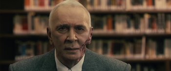 Movie still from “The Box” (2009), directed by Richard Kelly – An old man with a fake face and a book shelf in the background; Close Up shot, Over the shoulder angle