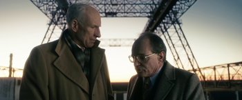 Movie still from “The Box” (2009), directed by Richard Kelly – Two older men standing next to each other in front of a metal structure; Medium shot, Low angle