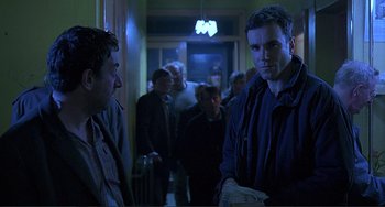 Movie still from “The Boxer” (1997), directed by Jim Sheridan – A man standing next to a group of people in a room; Medium shot, Over the shoulder angle