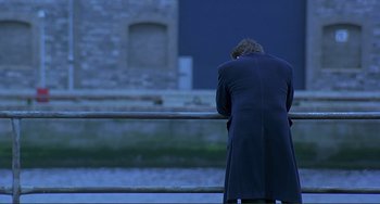 Movie still from “The Boxer” (1997), directed by Jim Sheridan – A man in a long coat standing on a bridge; Wide shot, Over the shoulder angle
