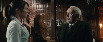 Movie still from “The Boy” (2016), directed by William Brent Bell – An older man wearing a suit and tie in front of a staircase; Medium shot, Over the shoulder angle