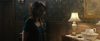 Movie still from “The Boy” (2016), directed by William Brent Bell – A woman standing next to a table in a room; Medium shot, Over the shoulder angle