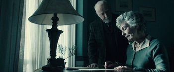 Movie still from “The Boy” (2016), directed by William Brent Bell – An older man and a younger woman looking at papers on a desk; Medium shot, Over the shoulder angle