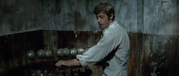 Movie still from “The Brain” (1969), directed by Gérard Oury – A man in a white shirt is standing in front of a bunch of cans; Medium shot, Low angle