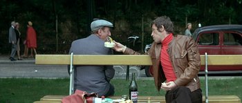 Movie still from “The Brain” (1969), directed by Gérard Oury – Two men sitting on a bench eating food; Medium shot, High angle