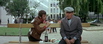 Movie still from “The Brain” (1969), directed by Gérard Oury – Two men are sitting on a bench and one of them is eating a sandwich; Medium shot, High angle