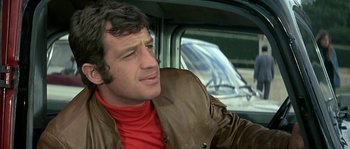 Movie still from “The Brain” (1969), directed by Gérard Oury – A person sitting in a car wearing a leather jacket; Close Up shot, Low angle