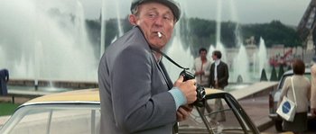 Movie still from “The Brain” (1969), directed by Gérard Oury – An older man holding a camera while smoking a cigarette; Medium shot, Low angle